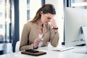 A stressed woman at her desk, holding glasses and pinching her nose in fatigue or tension.